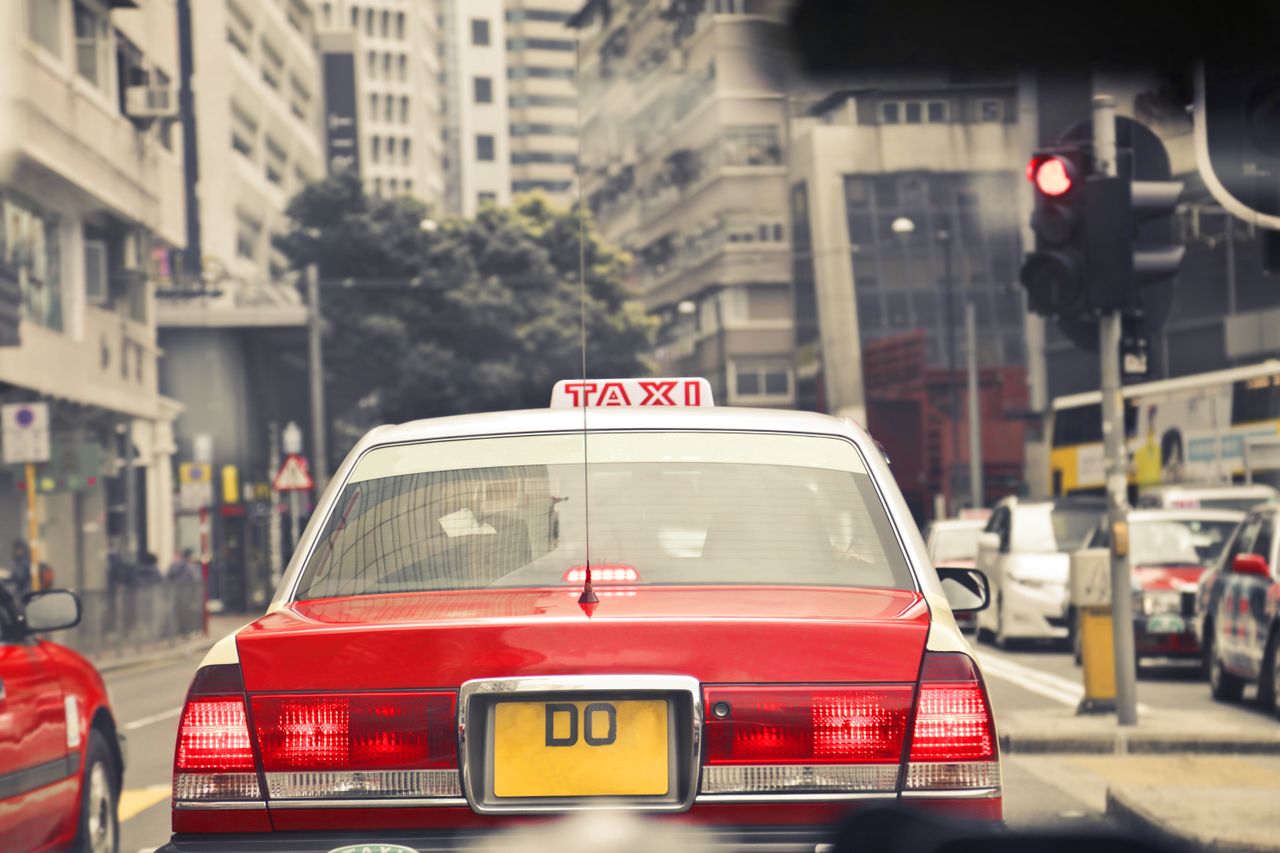 Red taxi amidst city traffic on a bustling Hong Kong street, under bright daylight.